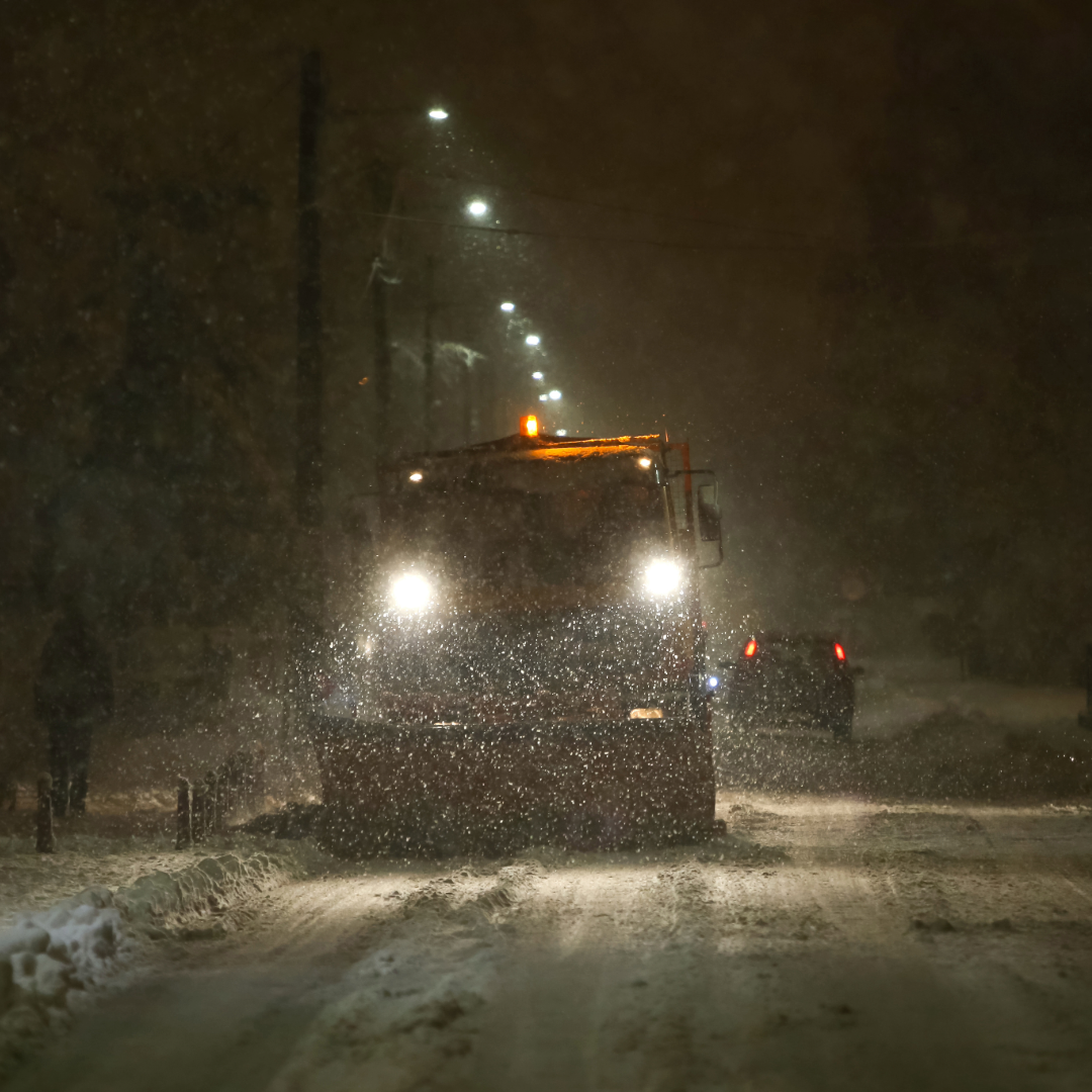 A heavy-duty snow plow truck clearing a thick layer of snow from a city street at night during a winter storm under glowing street lights.